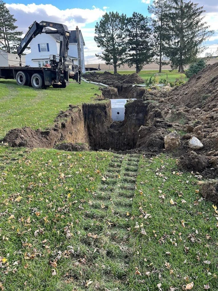 Boom truck setting a concrete distribution box into a deep trench beside a farmhouse, with the yard already restored and seeded in the foreground